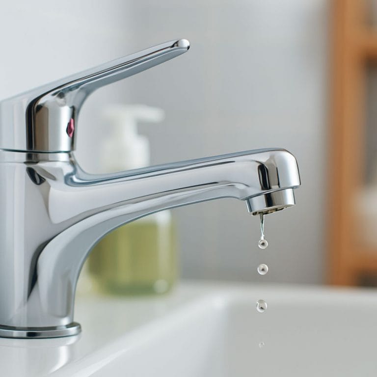 A chrome faucet dripping water into a bathroom sink, showing a minor leak with droplets falling from the spout.