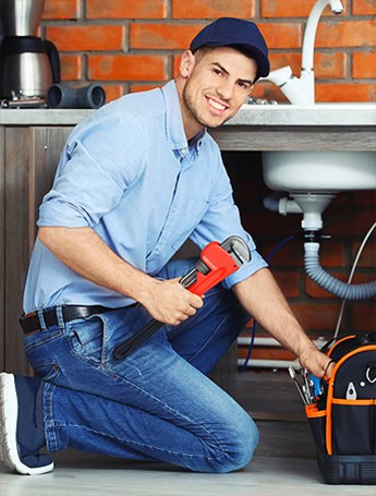A plumber kneels under a kitchen sink with a wrench and tool bag while troubleshooting plumbing issues around the drain pipes.